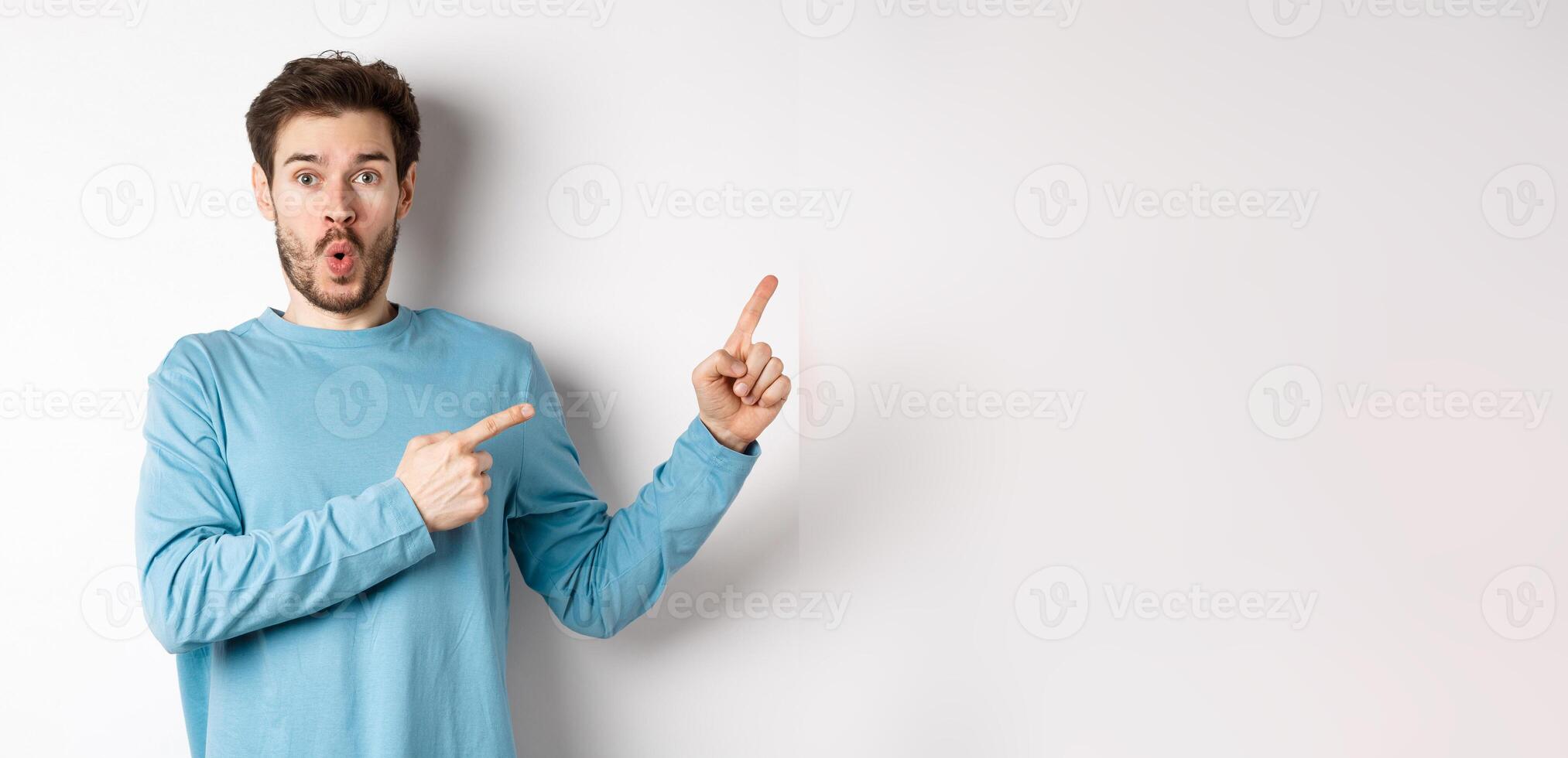 Wow look here. Impressed young man pointing fingers right and showing logo, standing amazed on white background photo