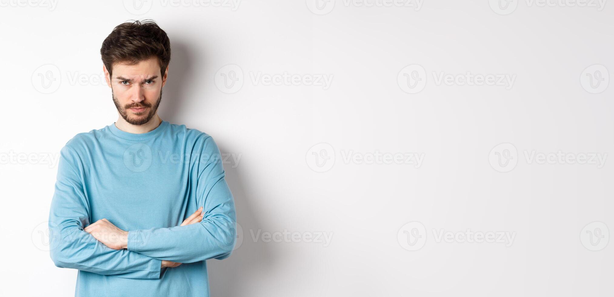 Image of offended and sad young man with beard, look from under forehead and sulking irritated, cross arms on chest defensive, mad at someone, standing on white background photo