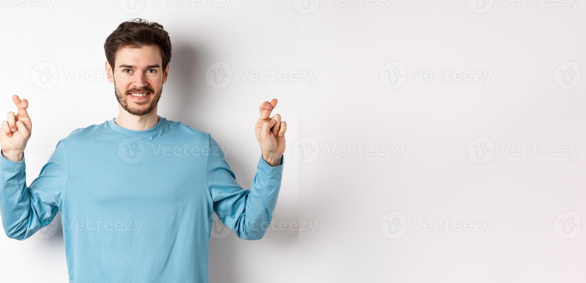Optimistic smiling man cross fingers for good luck, making wish and waiting for results, standing over white background photo