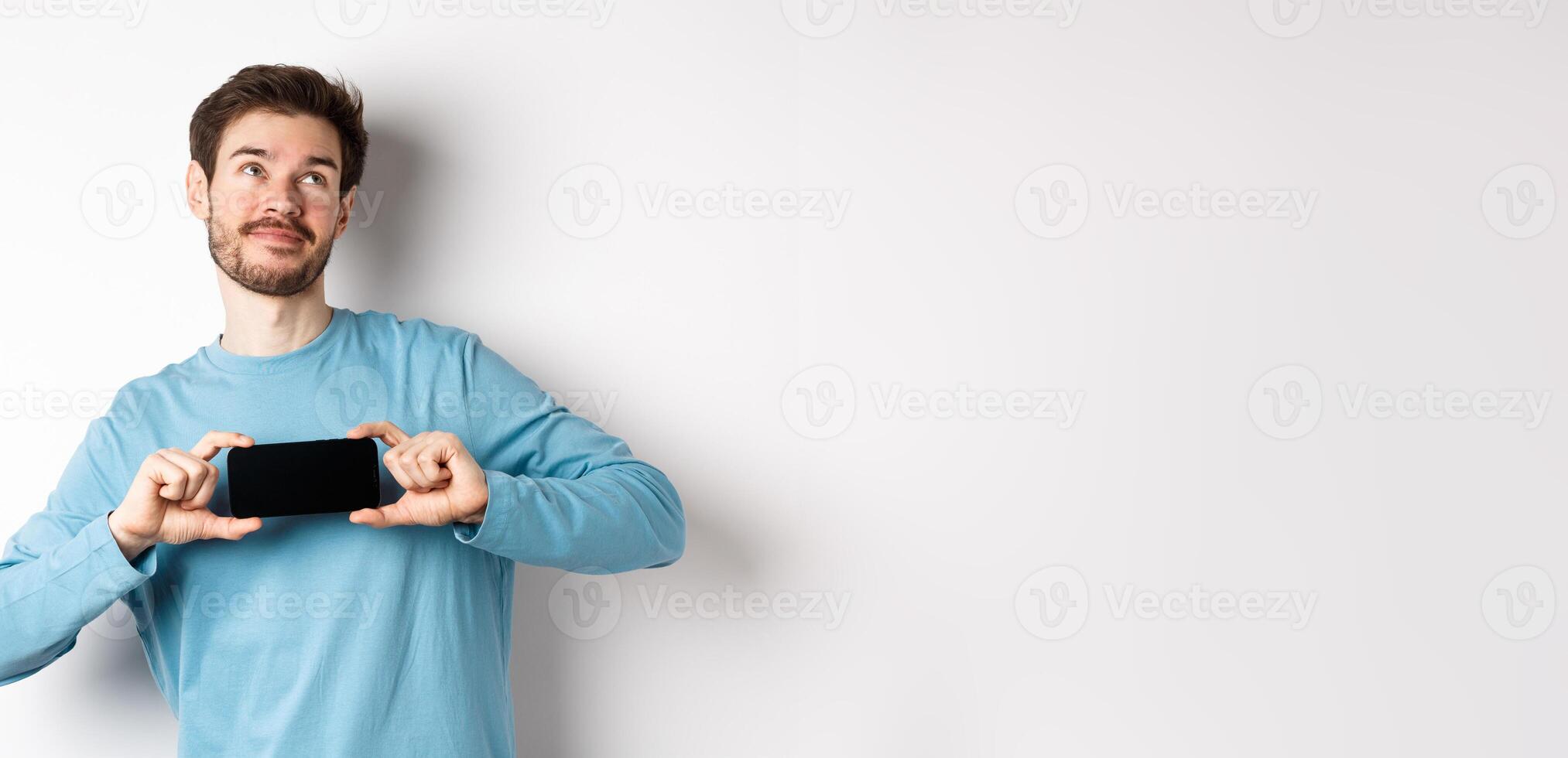 E-commerce and shopping concept. Smiling young man showing mobile screen and dreaming about something, looking at upper right corner nostalgic, standing over white background photo