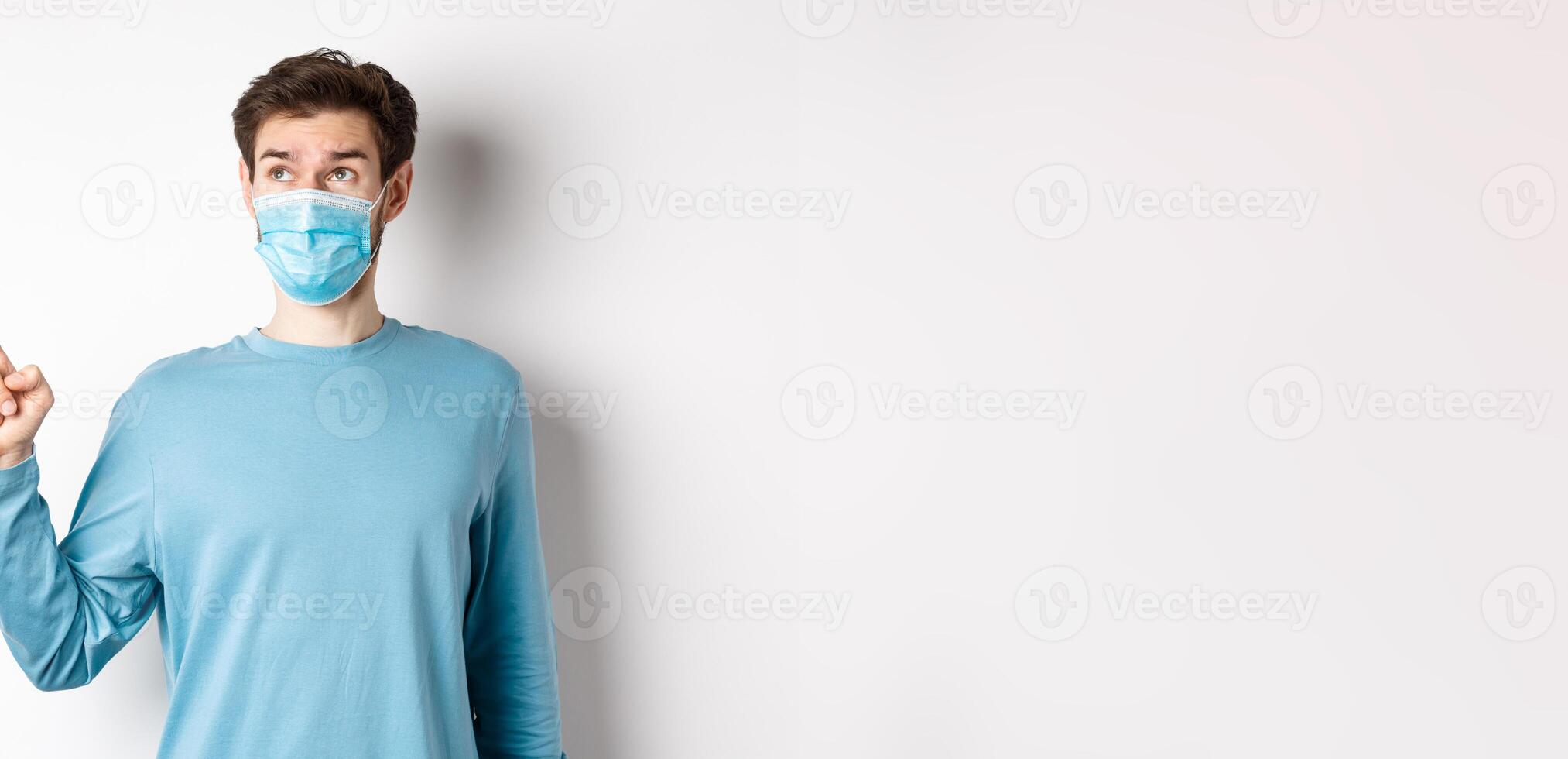 Coronavirus, health and quarantine concept. Curious guy in medical mask, pointing and looking at upper left corner banner, standing on white studio background photo
