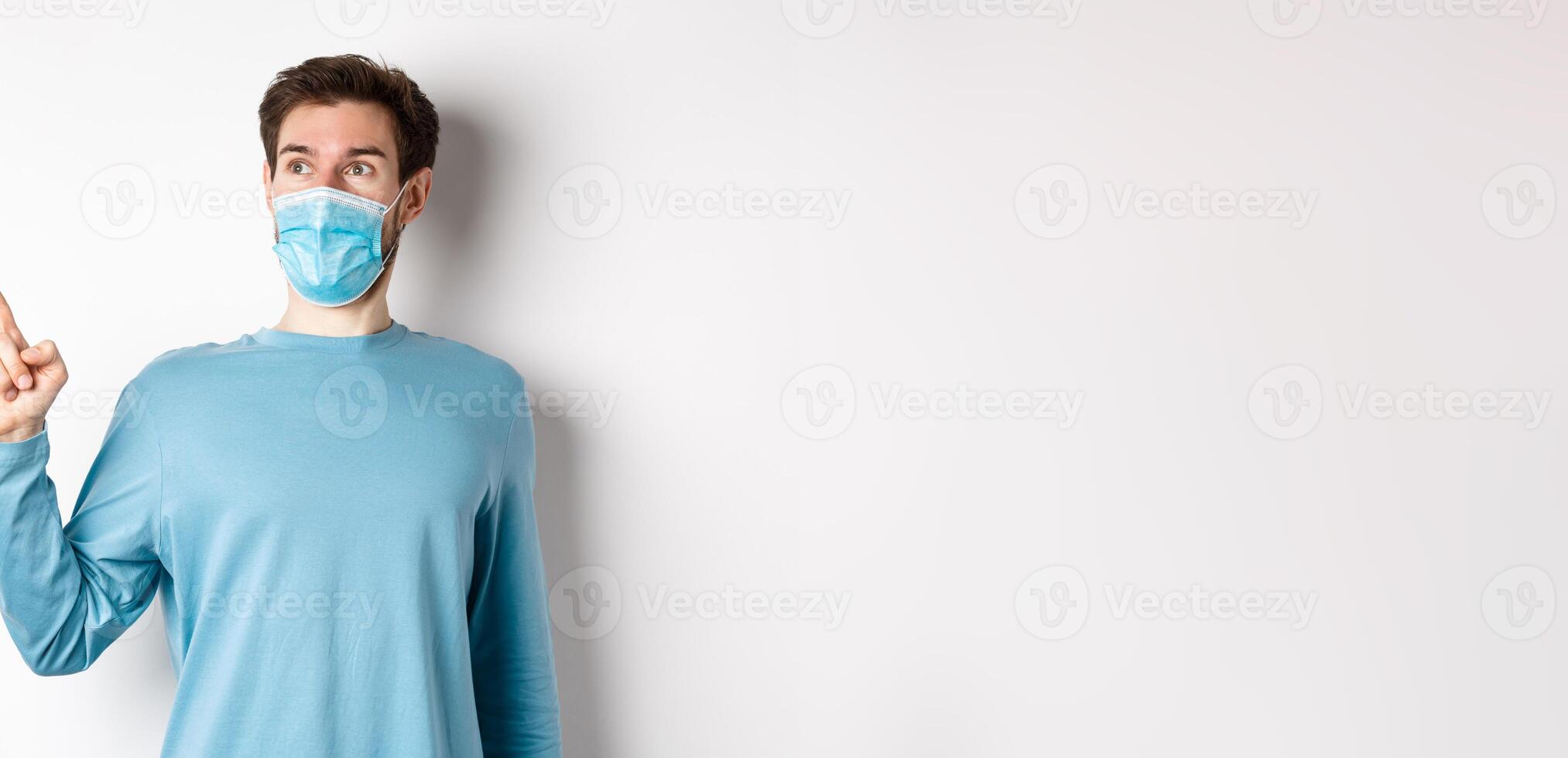 Coronavirus, health and quarantine concept. Surprised guy looking in awe and pointing at upper left corner, standing in medical mask and sweatshirt over white background photo