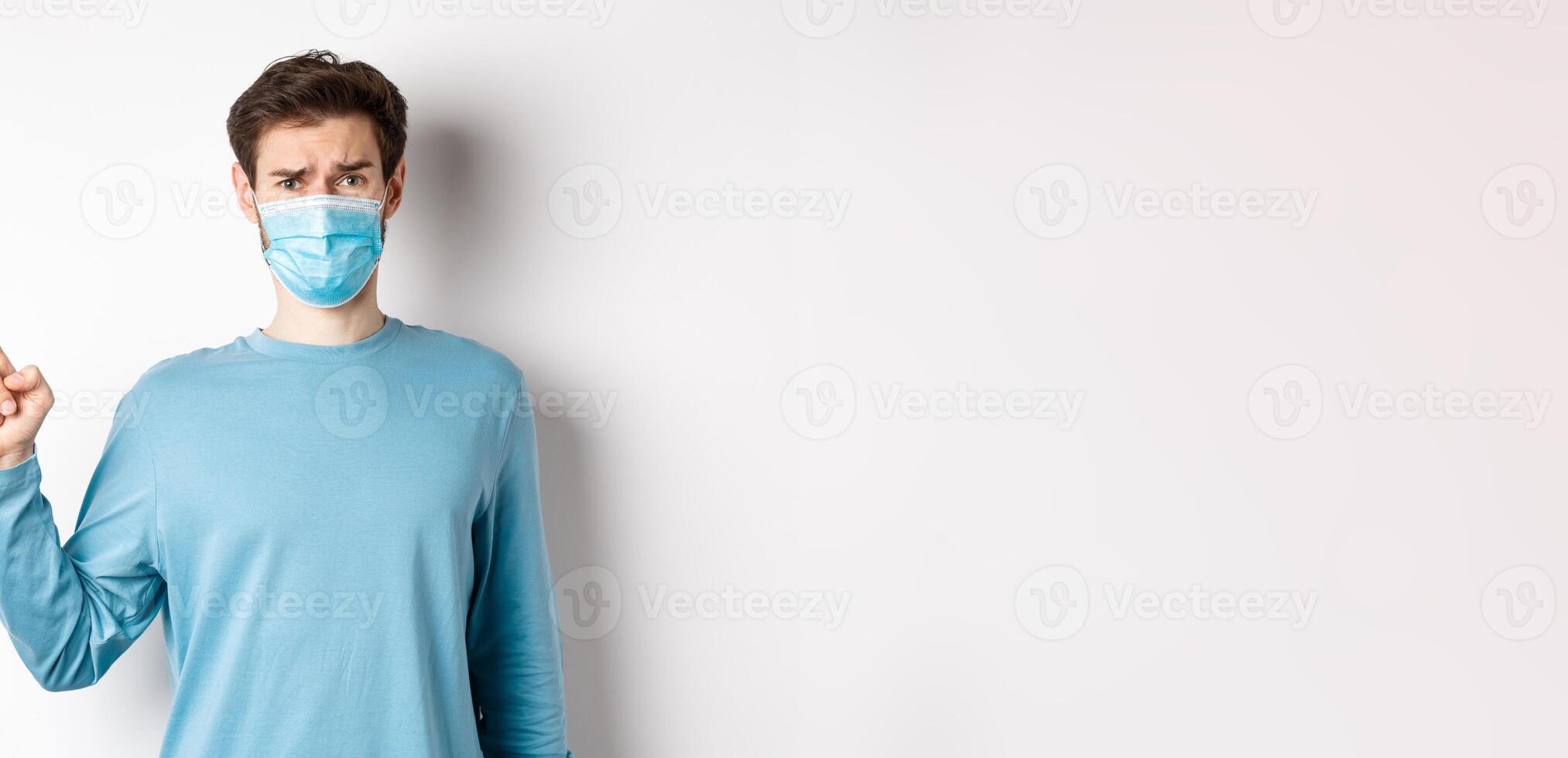 Coronavirus, health and quarantine concept. Skeptical man in medical mask pointing finger left at logo, frowning disappointed, standing over white background photo