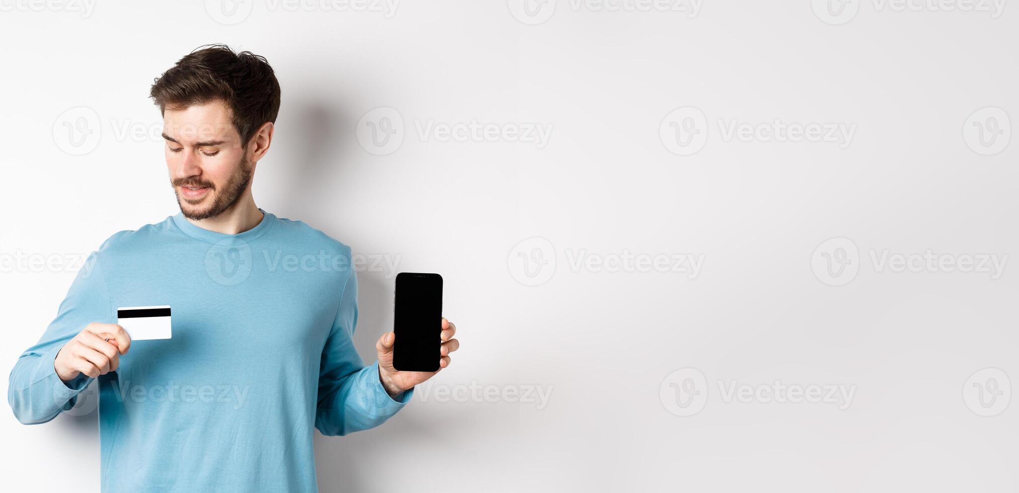 Handsome young man with beard, showing empty smartphone screen and looking at plastic credit card, standing over white background photo