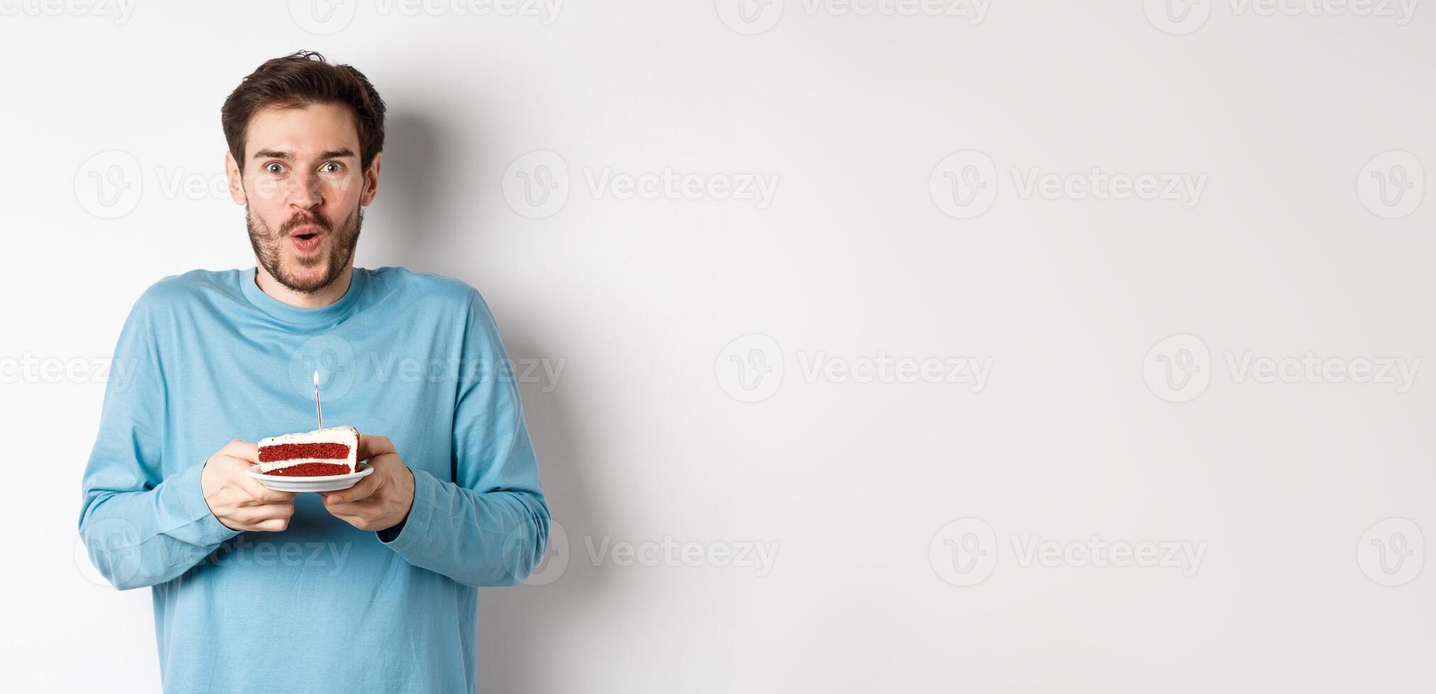 Excited young man making wish on birthday cake with lit candle, celebrating bday, standing on white background photo