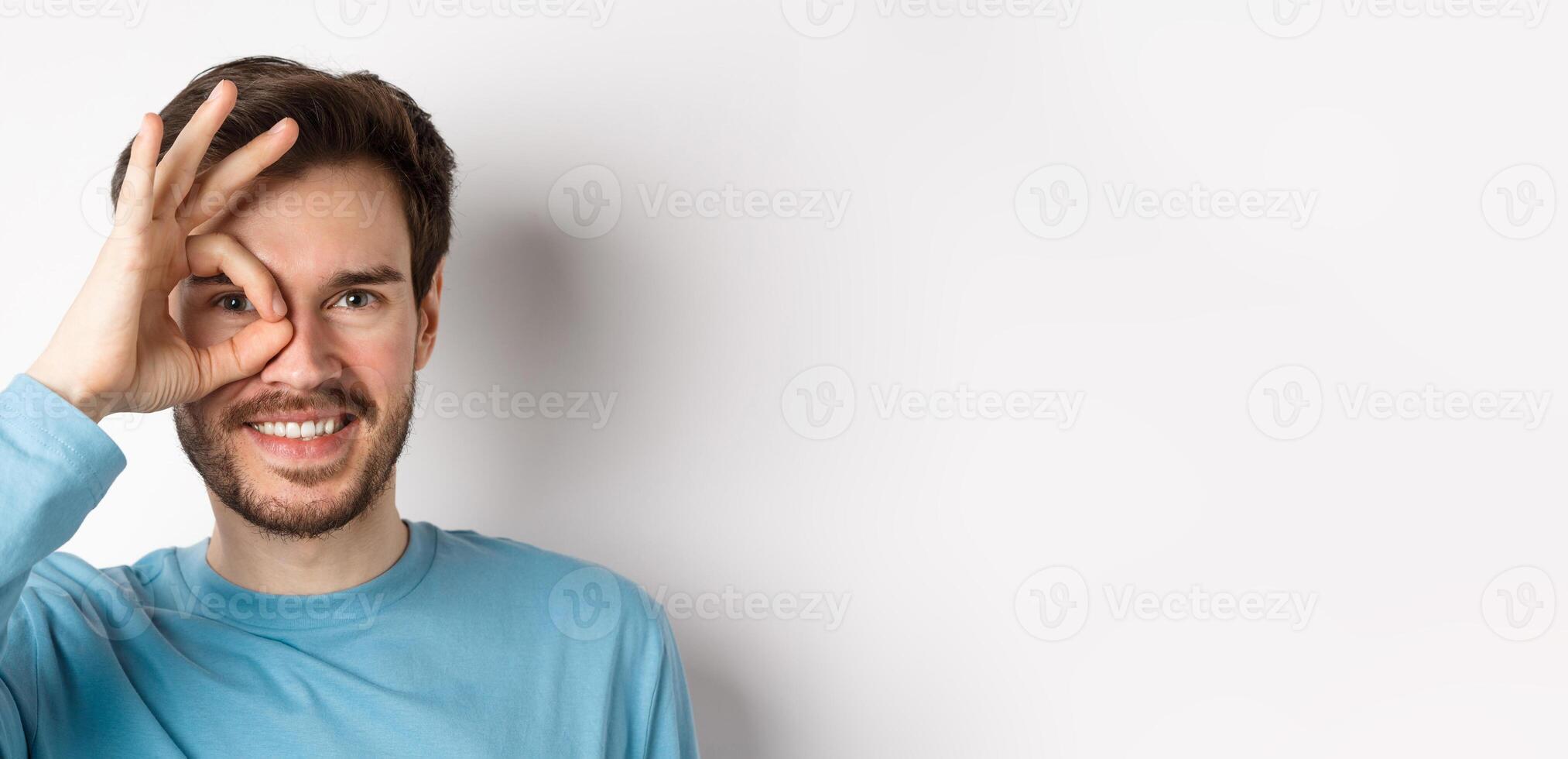 Close up of happy man looking through okay sign and smiling, approve and like something good, standing over white background photo