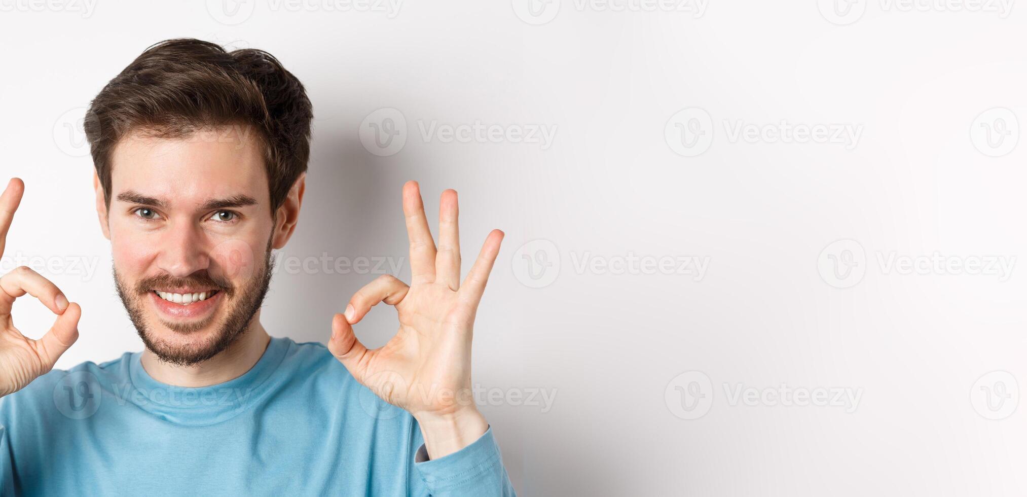 Close-up of confident young man smiling, showing okay signs, praising excellent choice, approve something good, standing over white background photo