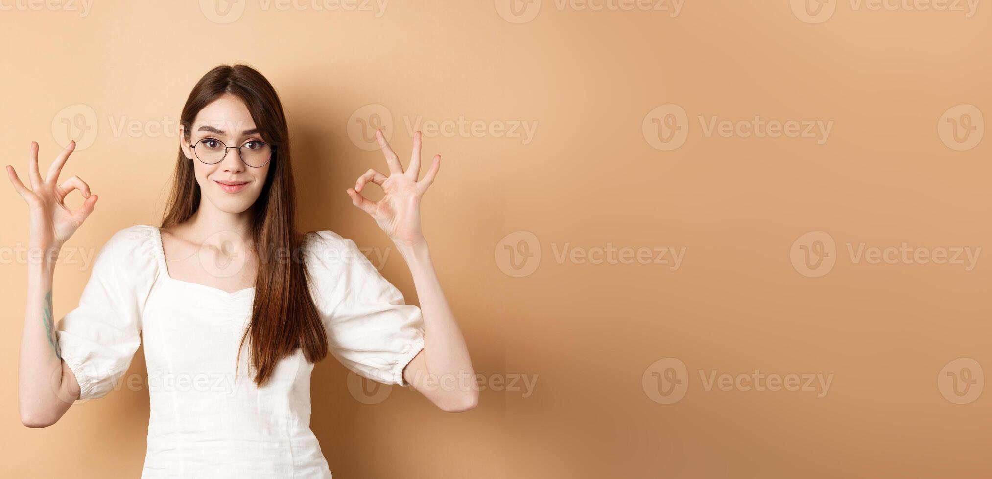 Satisfied female model in glasses show okay sign, looking pleased, agree and approve, standing on beige background photo