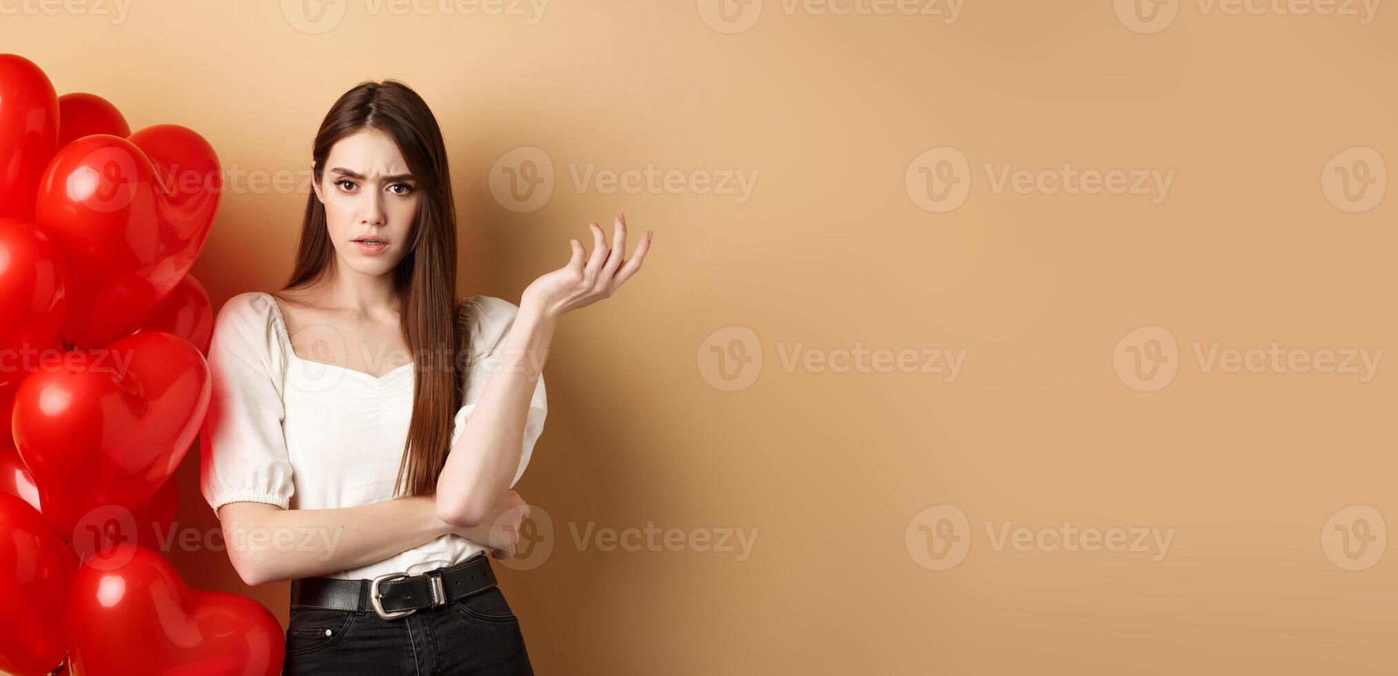 Valentines day and love concept. Confused girlfriend having argument with lover, raising hand and looking annoyed at camera, cant understand something, standing near hearts balloons photo
