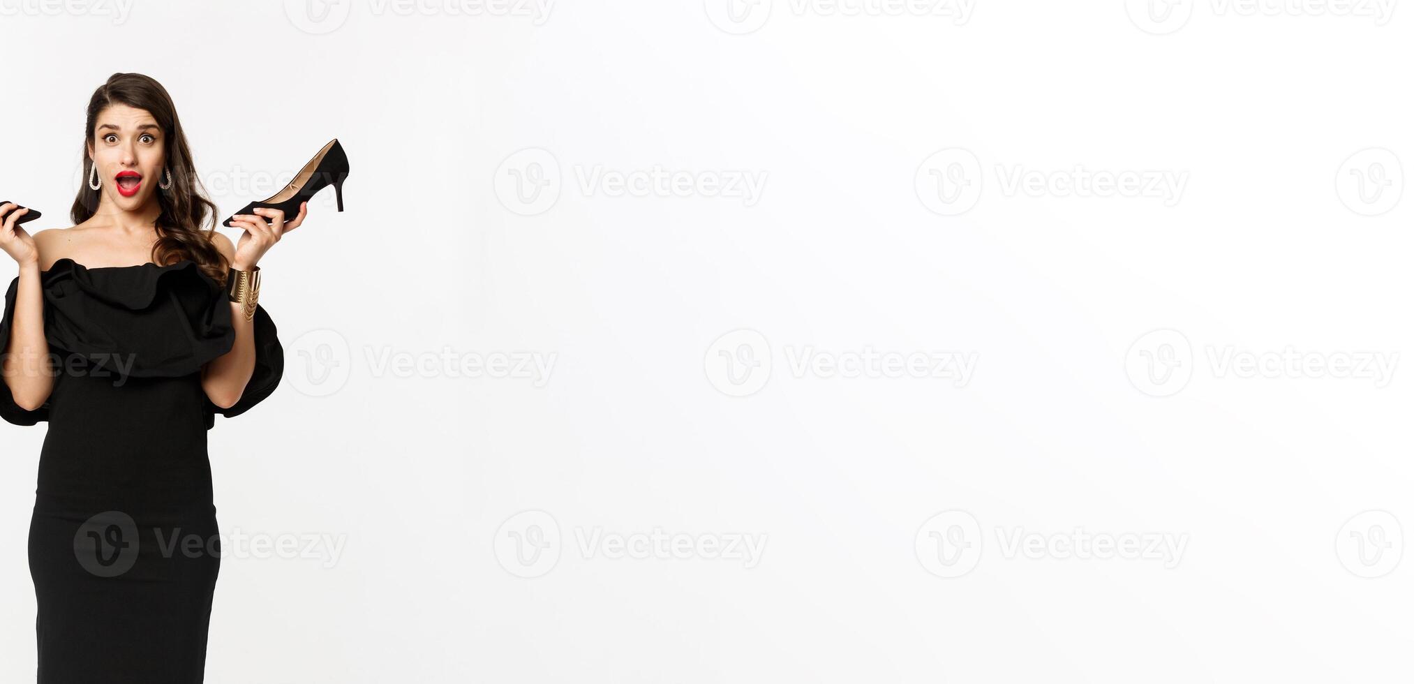 Beauty and fashion concept. Full length of excited glamour woman in black dress, showing high heels and looking excited, dressing up for party, white background photo