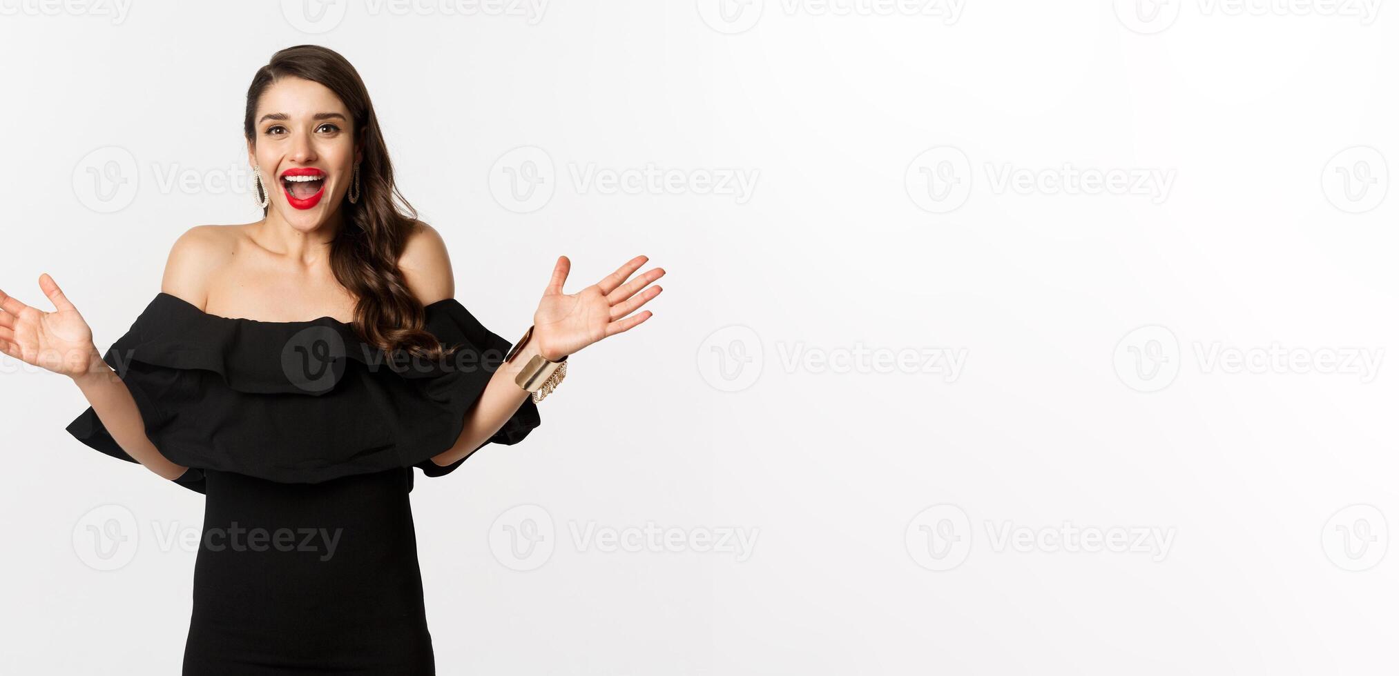 Beauty and fashion concept. Image of surprised and happy young woman in party dress reacting to good news, raising hands up and smiling amazed, white background photo