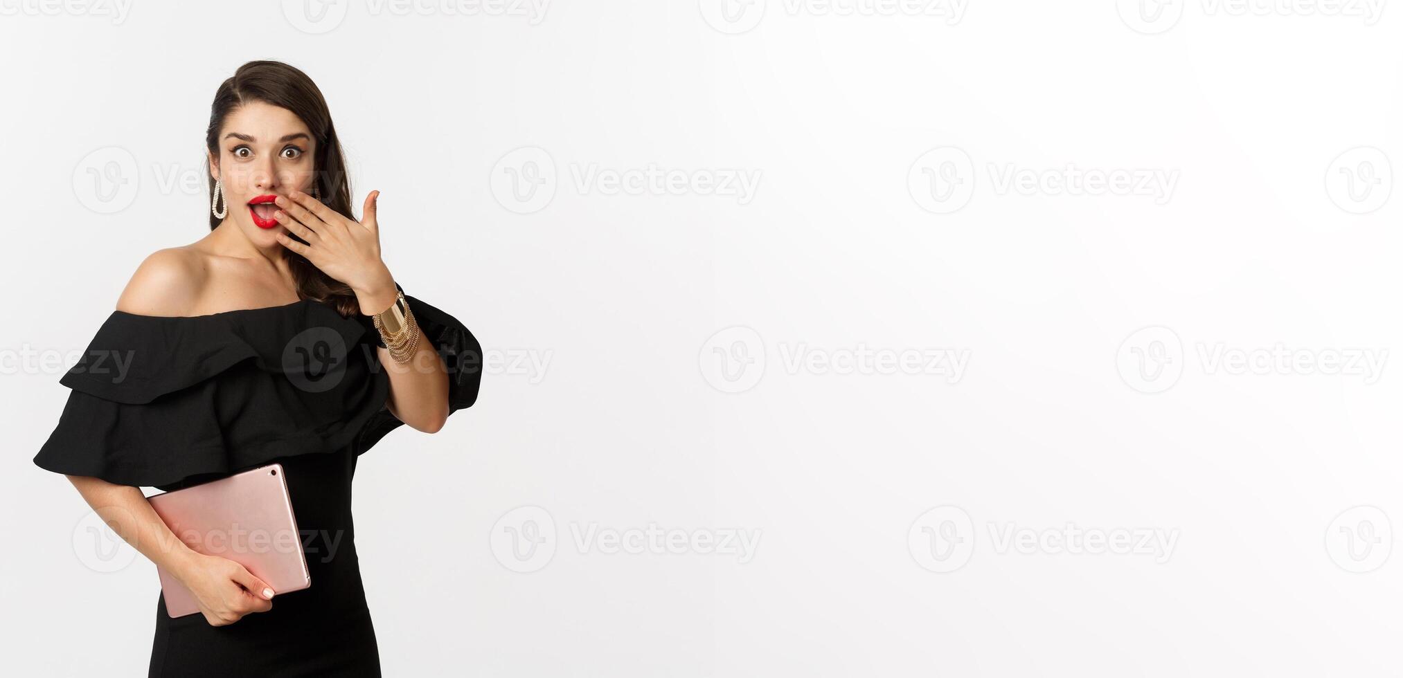 Fashion and shopping concept. Stylish young woman with glamour makeup, wearing black dress, holding digital tablet and looking surprised, white background photo