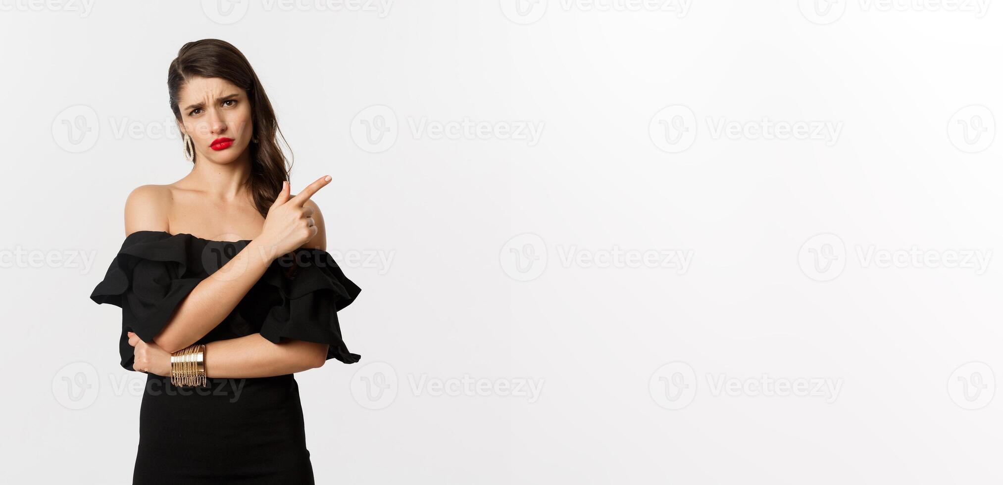 Fashion and beauty. Skeptical glamour woman with red lips, black dress, pointing finger right at something lame and boring, standing over white background photo