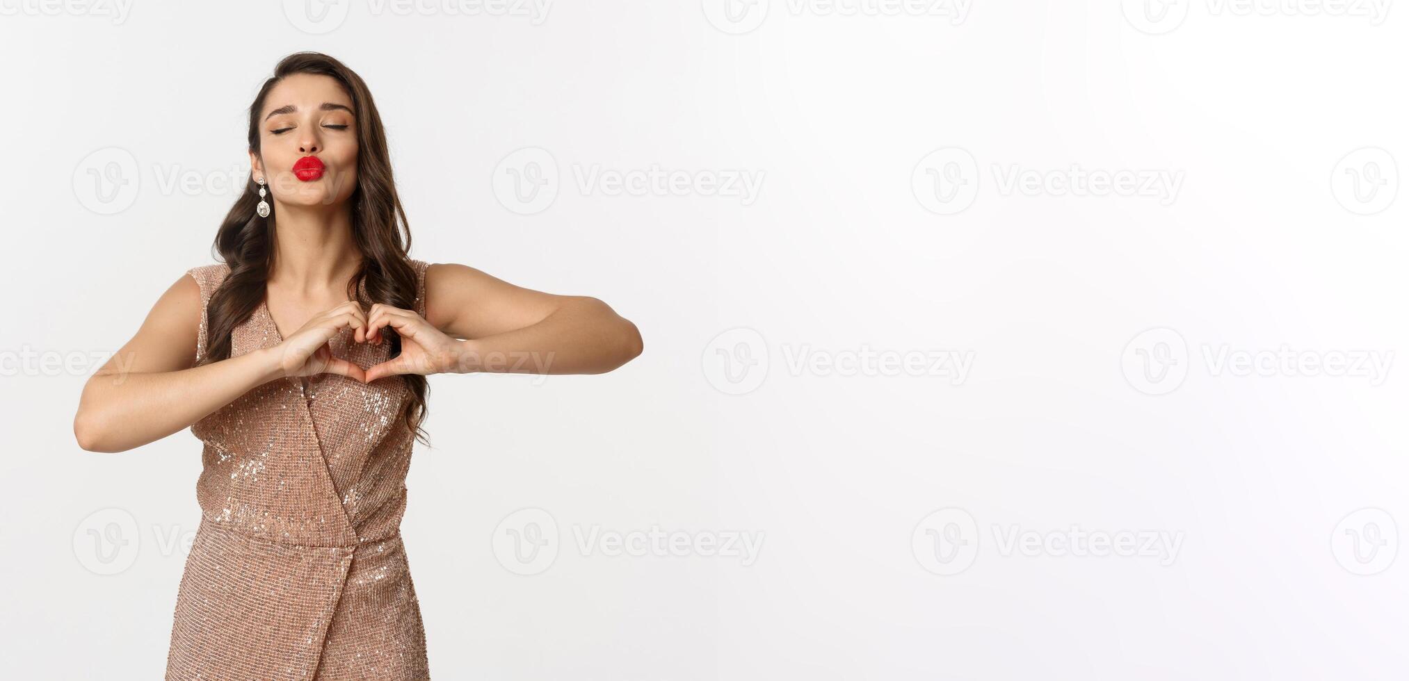 Celebration and party concept. Beautiful young woman in stylish dress, red lips, showing heart sign and waiting for kiss, standing over white background photo