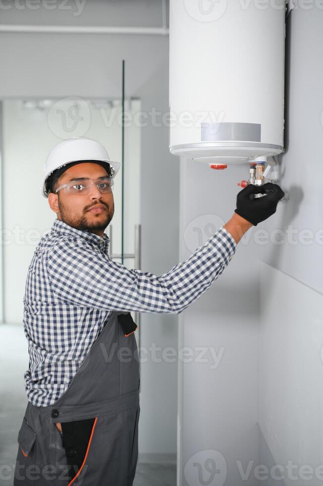 Indian plumber using screwdriver while fixing boiler or water heater, working on heating system in apartment photo