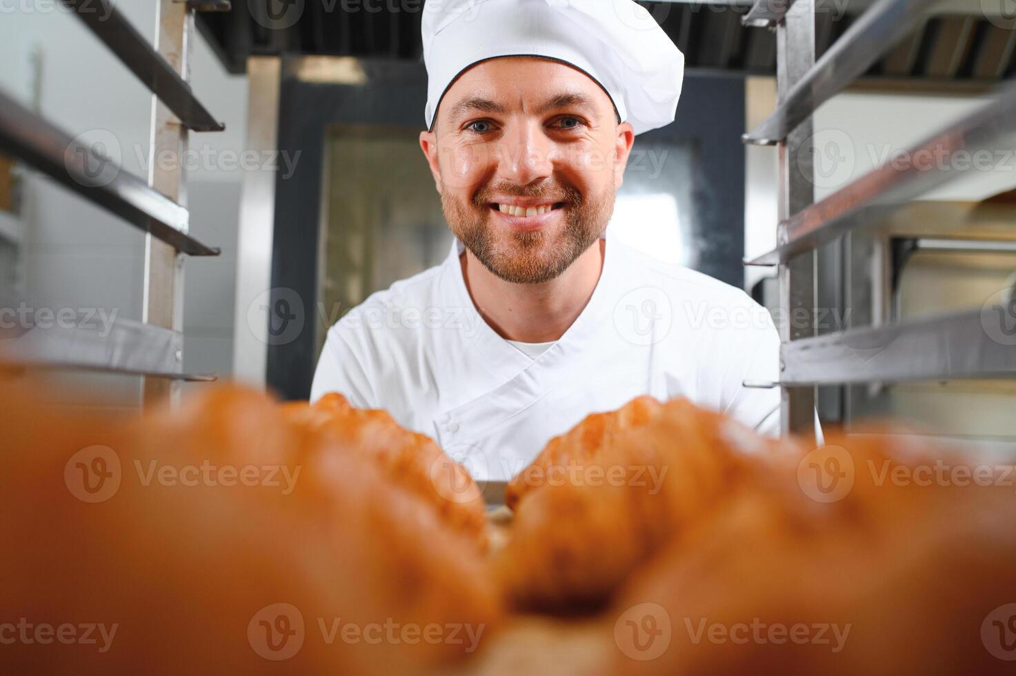 Handsome baker in uniform holding tray full of freshly baked croissants at the manufacturing photo