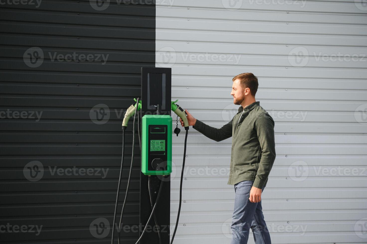 handsome bearded man in casual wear, standing at the charging station and charger for an electric car. Eco electric car concept photo