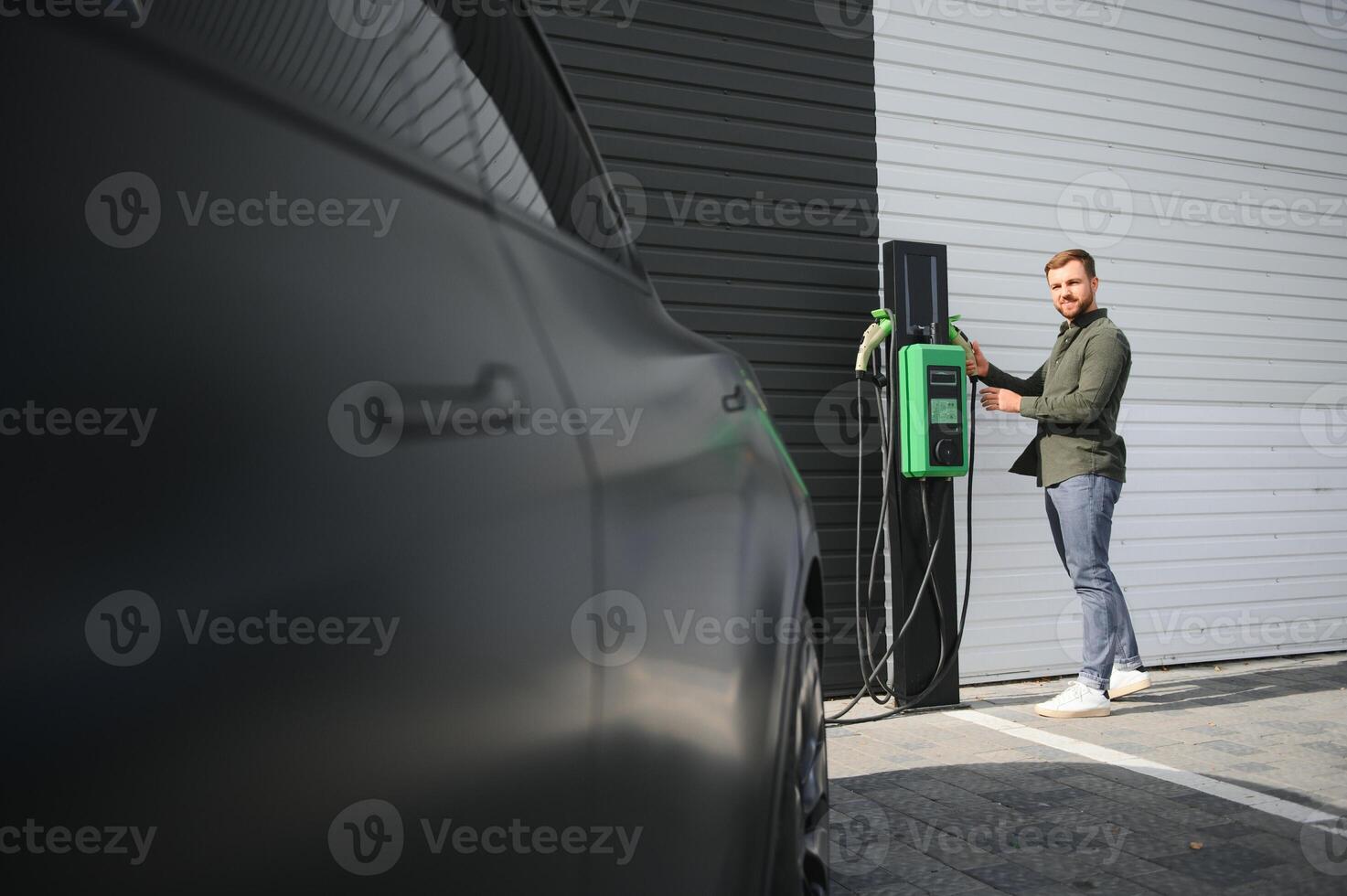 handsome bearded man in casual wear, standing at the charging station and charger for an electric car. Eco electric car concept photo