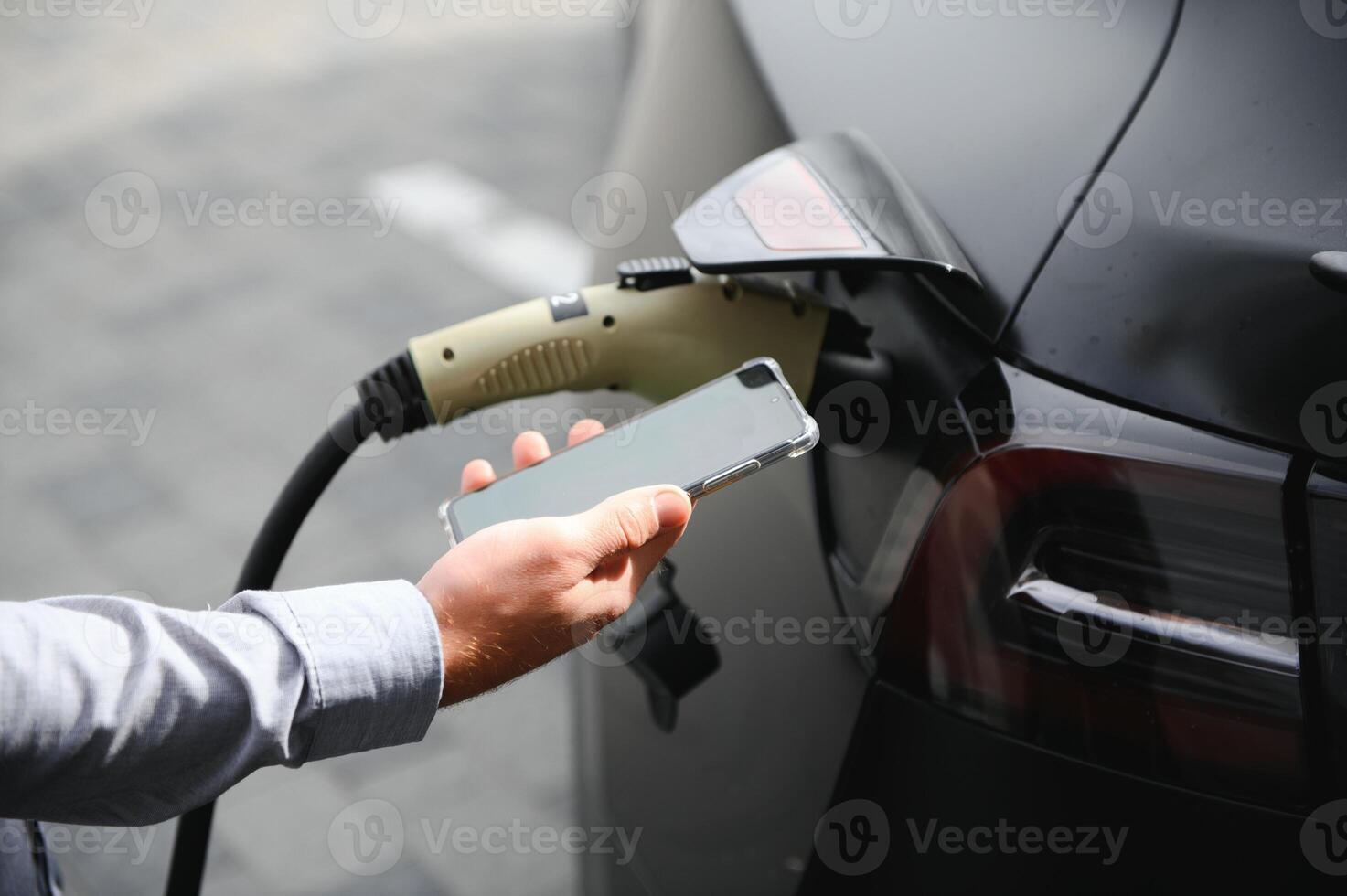 A man charges an electric car and checks the charge level on a smartphone photo