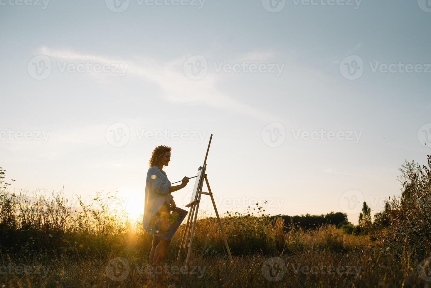 Silhouette of a blonde girl artist. Lady paints a painting on the canvas with the help of paints. A wooden easel keeps the picture. Summer is a sunny day, sunset. photo