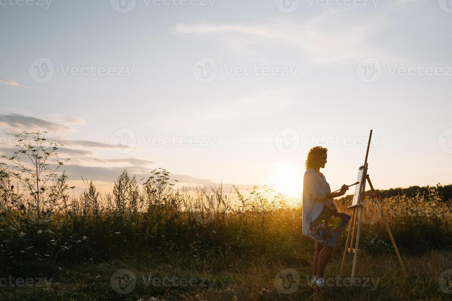 Silhouette of a blonde girl artist. Lady paints a painting on the canvas with the help of paints. A wooden easel keeps the picture. Summer is a sunny day, sunset. photo