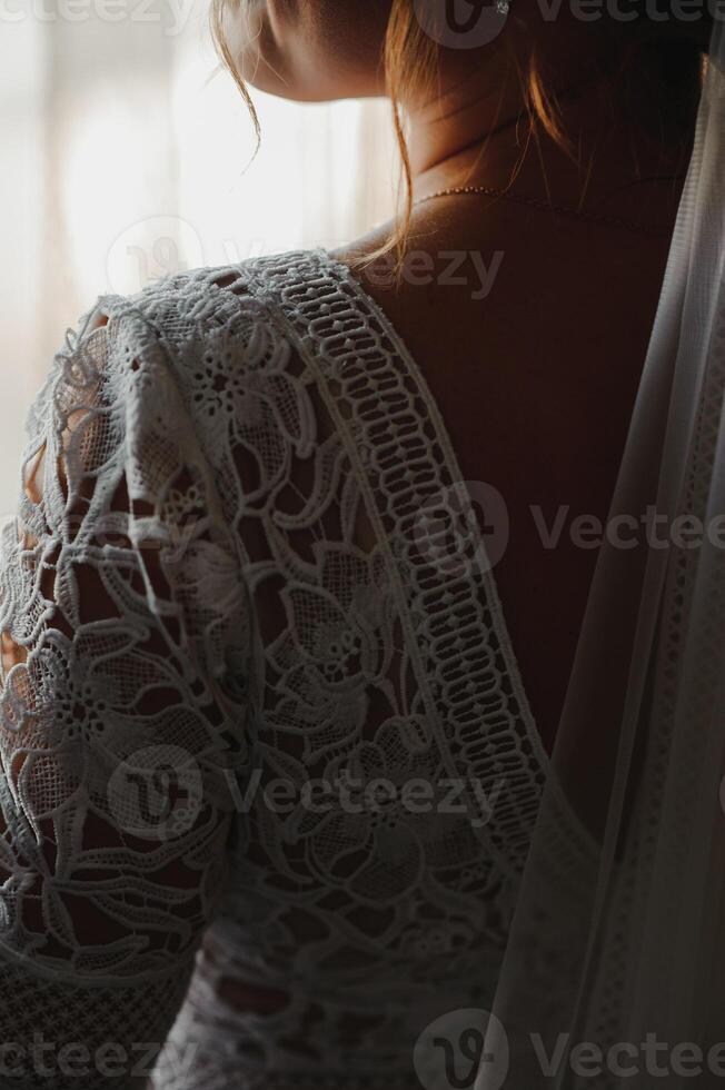Bride in a apartment in a wedding dress. Bride looks out of the window, wedding day. Charming bride stand in front of window in a white wedding dress. Girl stand by the window at her room. photo