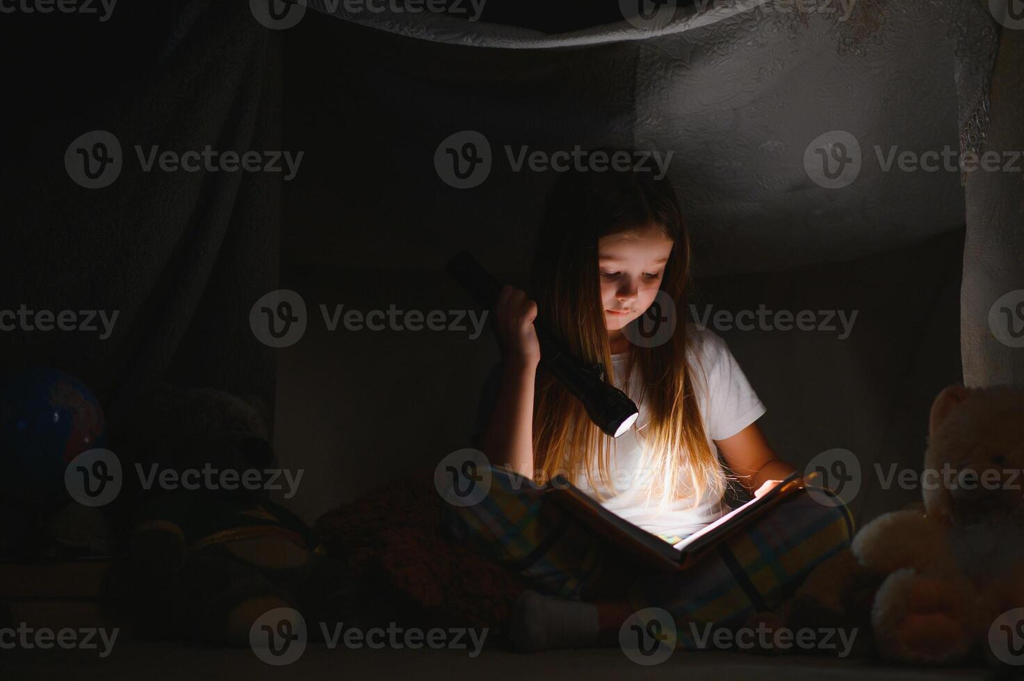 child girl reading with book and flashlight and teddy bear in tent. before going to bed photo