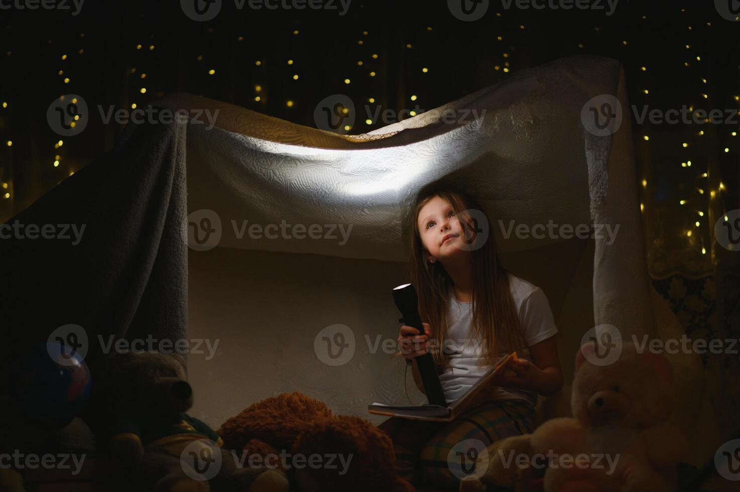 child girl reading with book and flashlight and teddy bear in tent. before going to bed photo