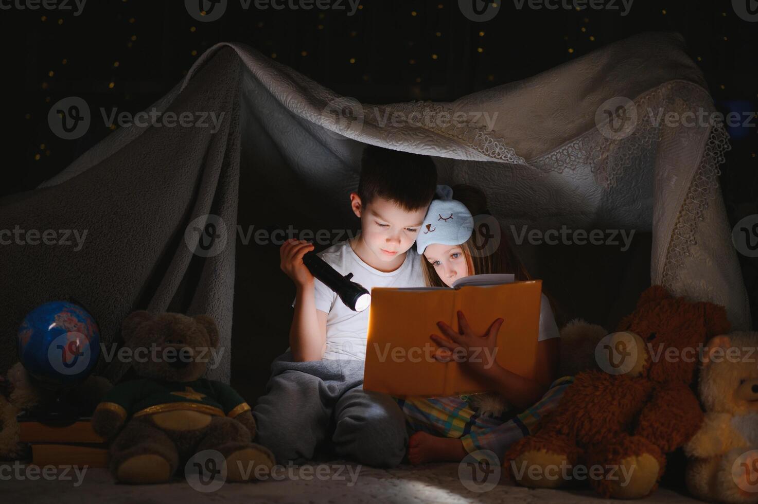 Two children with flashlight read a book under a blanket as a tent photo