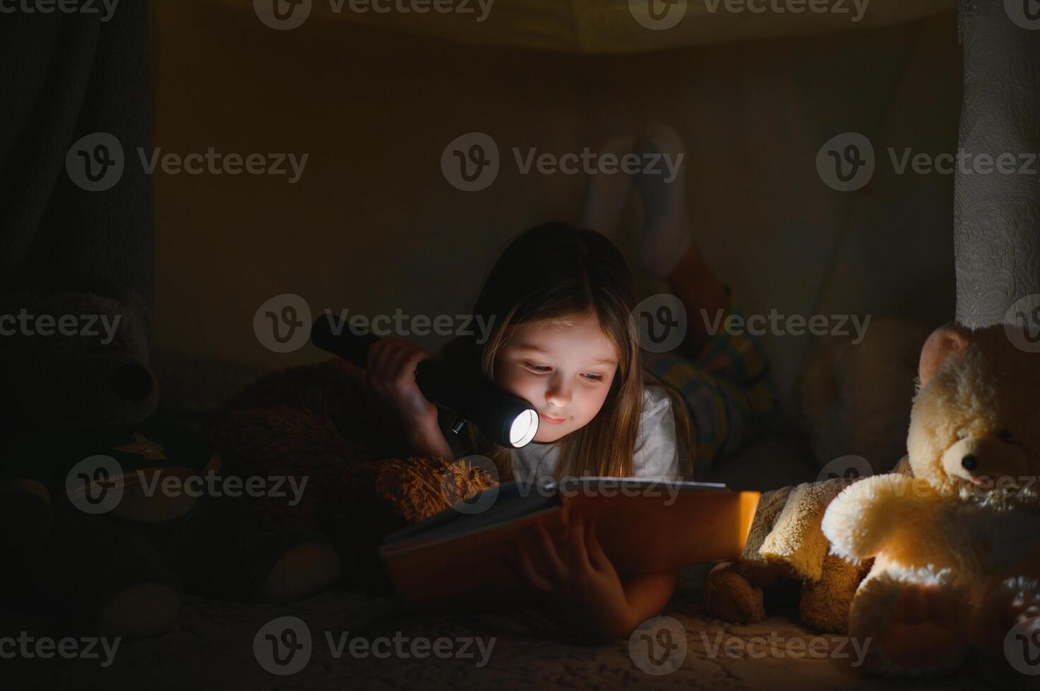 child girl reading with book and flashlight and teddy bear in tent. before going to bed photo