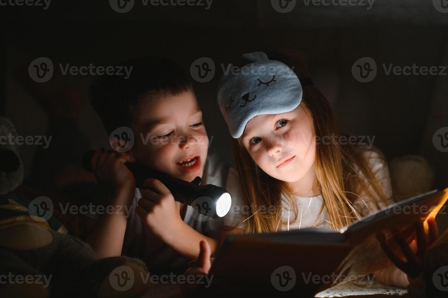 Two children with flashlight read a book under a blanket as a tent photo
