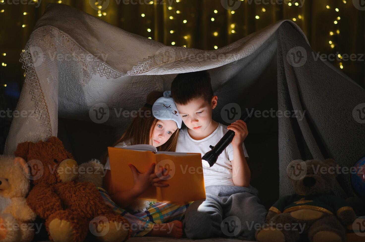 Two children with flashlight read a book under a blanket as a tent photo