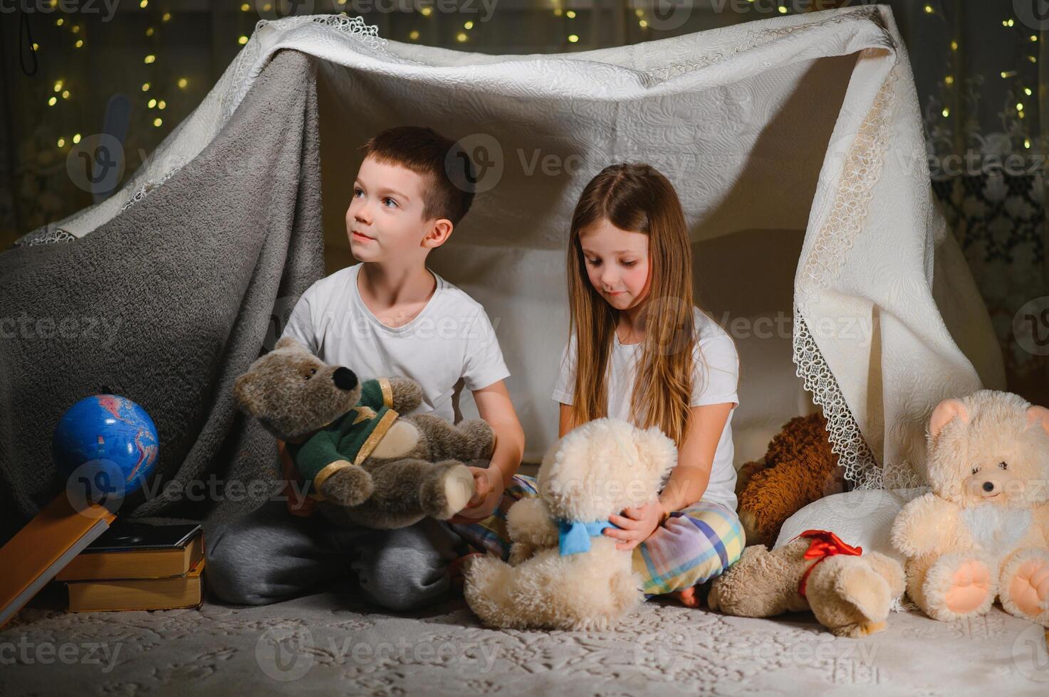 Two little child play at home in the evening to build a camping tent to read books with a flashlight and sleep inside. Concept of game, magic, creativity, alarm systems photo