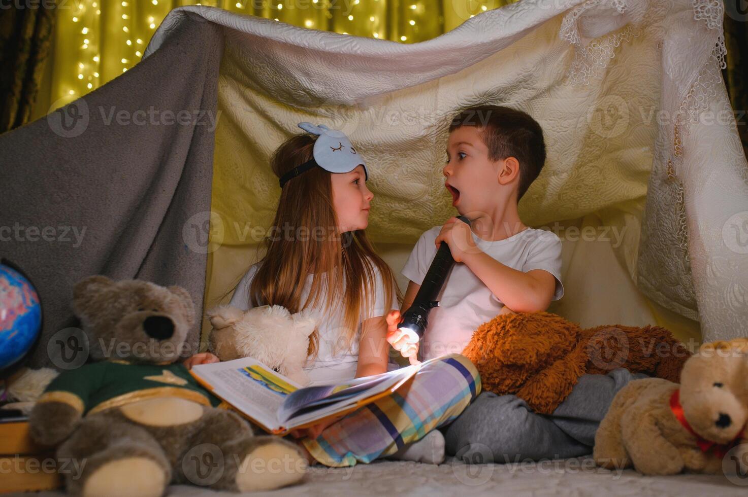 Two children with flashlight read a book under a blanket as a tent photo
