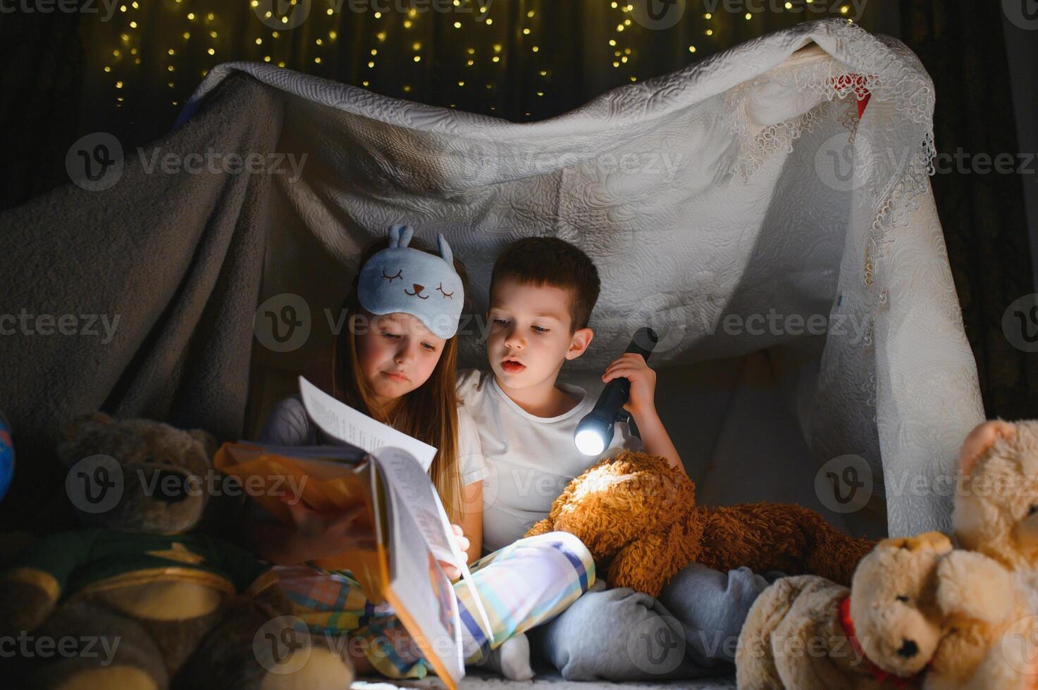 Little kids involving in reading amazing book. They lying in nice toy tent in playroom. Boy holding flashlight in hand photo