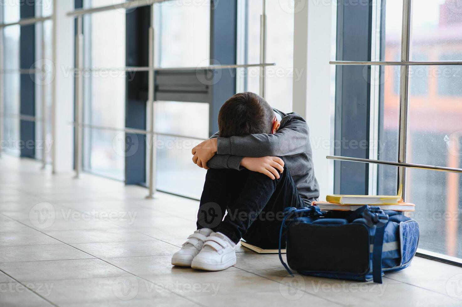 Little boy sitting alone on floor after suffering an act of bullying while children run in the background. Sad young schoolboy sitting on corridor with hands on knees and head between his legs photo