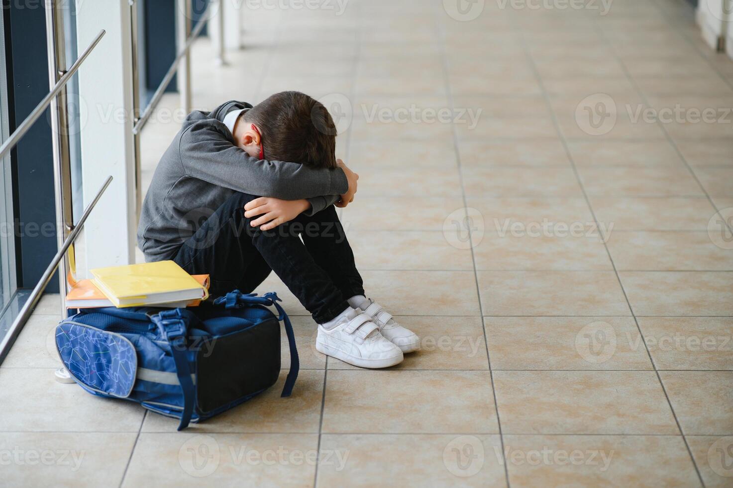 Little boy sitting alone on floor after suffering an act of bullying while children run in the background. Sad young schoolboy sitting on corridor with hands on knees and head between his legs photo