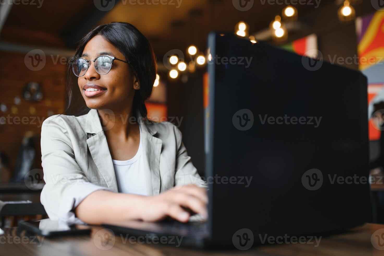 Beautiful african business woman waiting for the meeting in a co-working space and sitting with the portable computer. Hipster girl is surfing the web on a laptop during lunch. photo