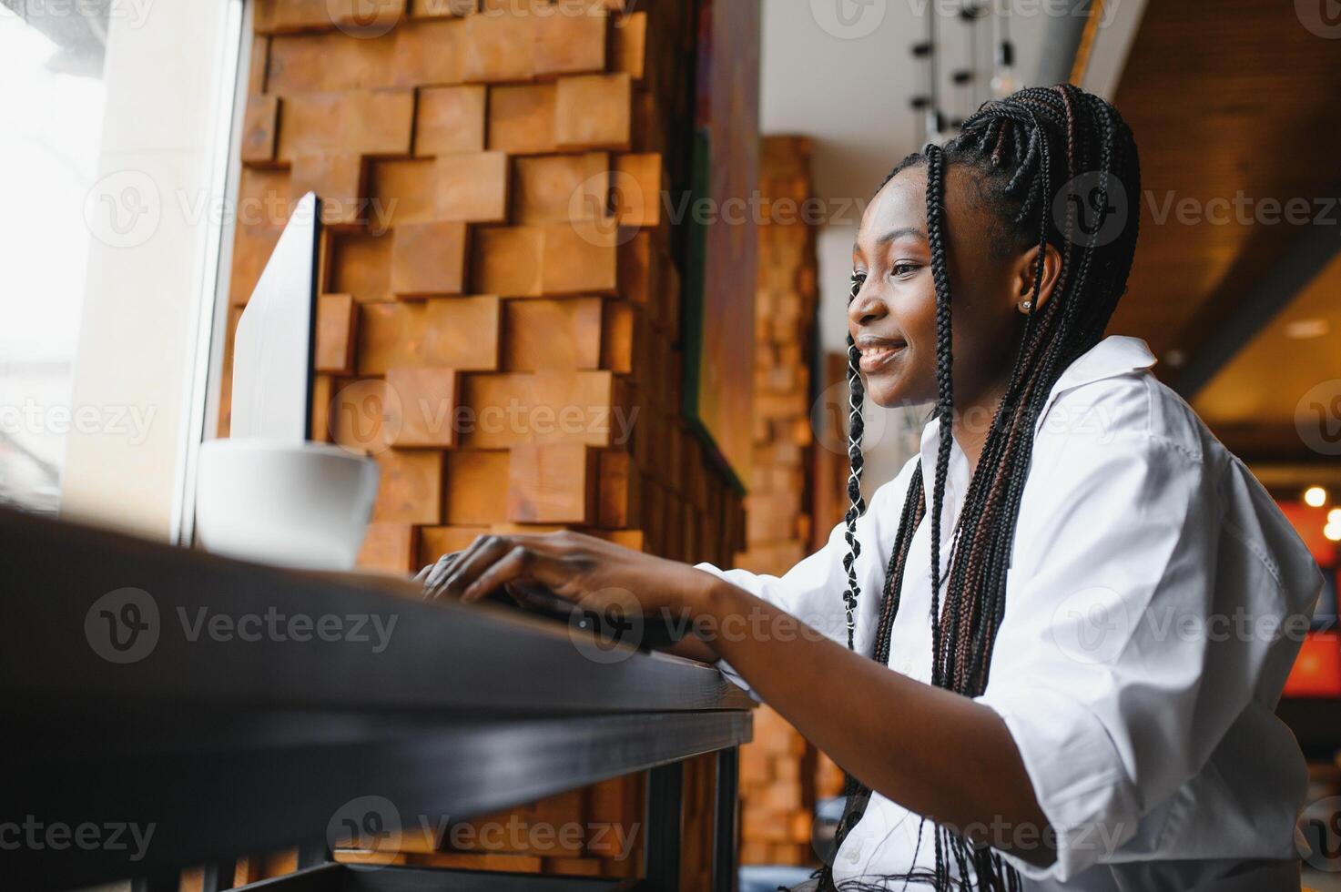Beautiful african business woman waiting for the meeting in a co-working space and sitting with the portable computer. Hipster girl is surfing the web on a laptop during lunch. photo
