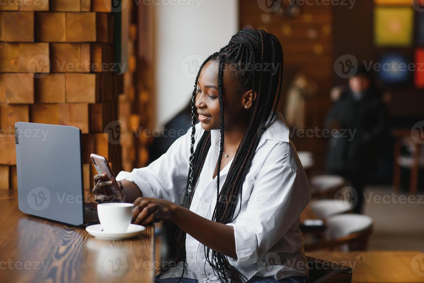 Beautiful african female with a long dark hair is typing messages on a smartphone while sitting beside the window in a soft light. Young business woman is using laptop and mobile phone for the work. photo