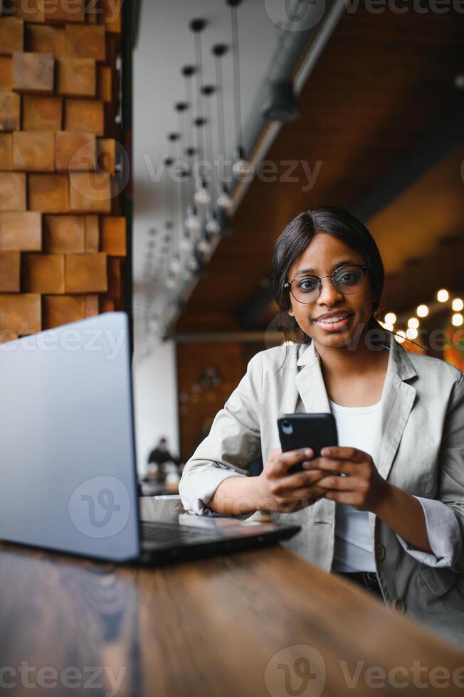 Beautiful african female with a long dark hair is typing messages on a smartphone while sitting beside the window in a soft light. Young business woman is using laptop and mobile phone for the work. photo
