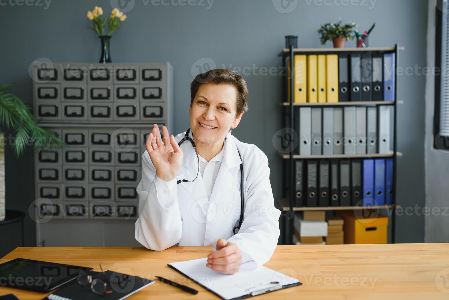 Portrait of female doctor counseling patient via call. Professional physician in white lab coat gesturing and explaining course of treatment sitting at office desk during online consultation. photo