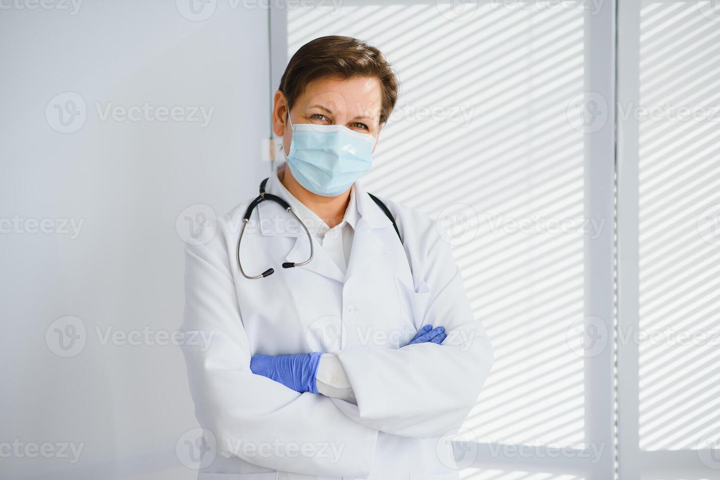 Older female doctor wearing face mask and white medical coat standing in hospital. Portrait photo
