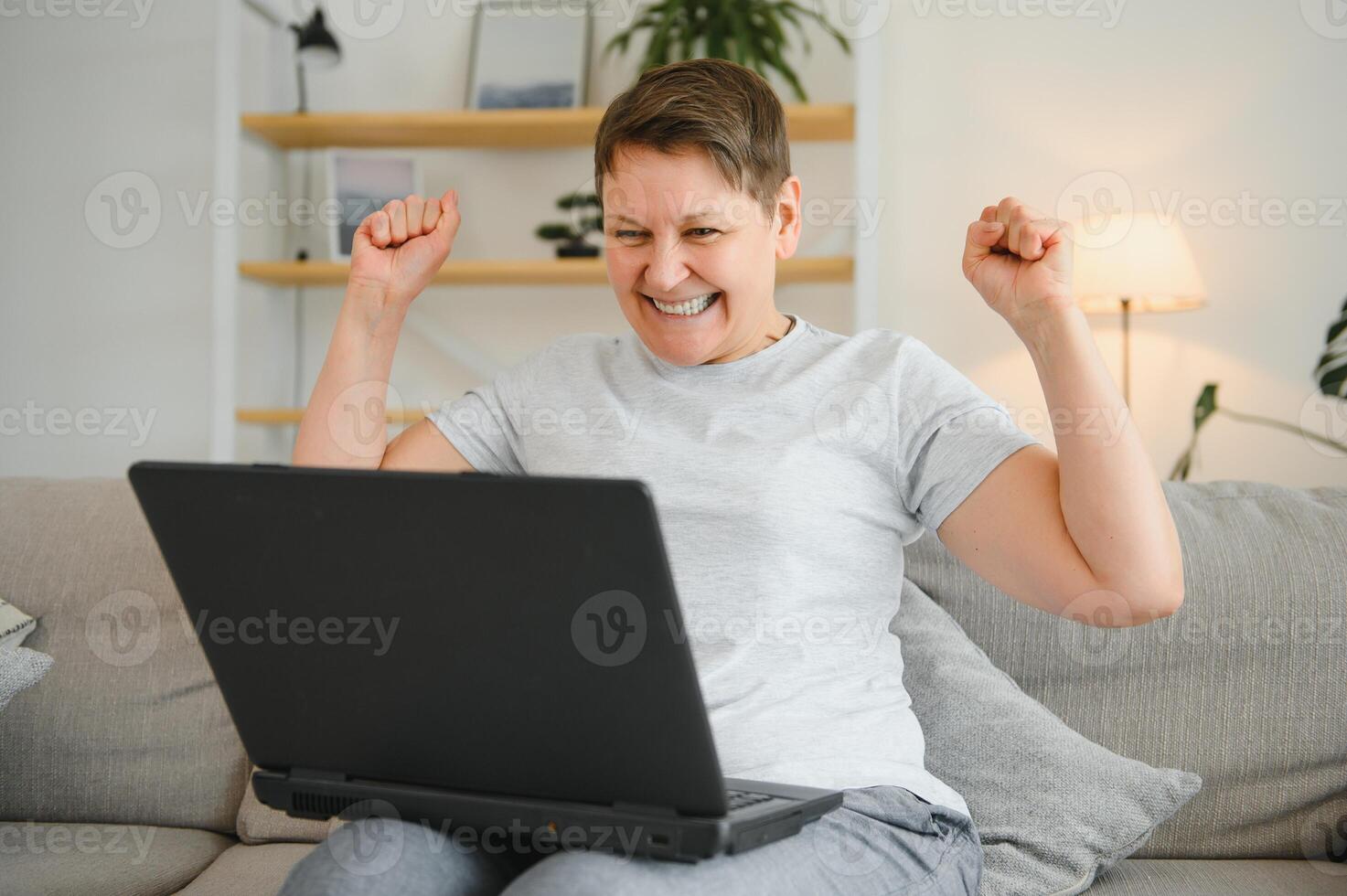 Excited mature woman looking at laptop screen, reading good news in message, celebrating online lottery win, rejoicing success, overjoyed older female sitting on couch at home, using computer. photo