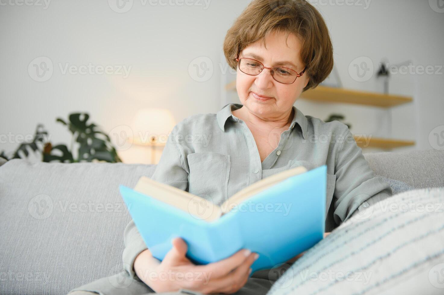 Restful reading. Portrait of thoughtful aged woman reading favorite literature at cozy home. She is lying on pillows on comfortable sofa. Legs are covered with soft blanket. photo