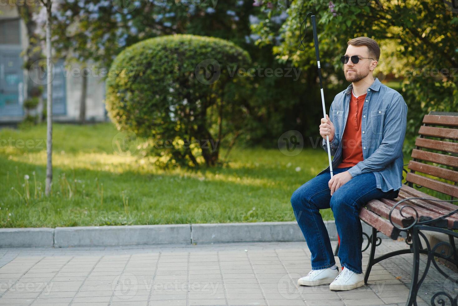 Visually impaired man with walking stick, sitting on bench in city park. Copy space photo