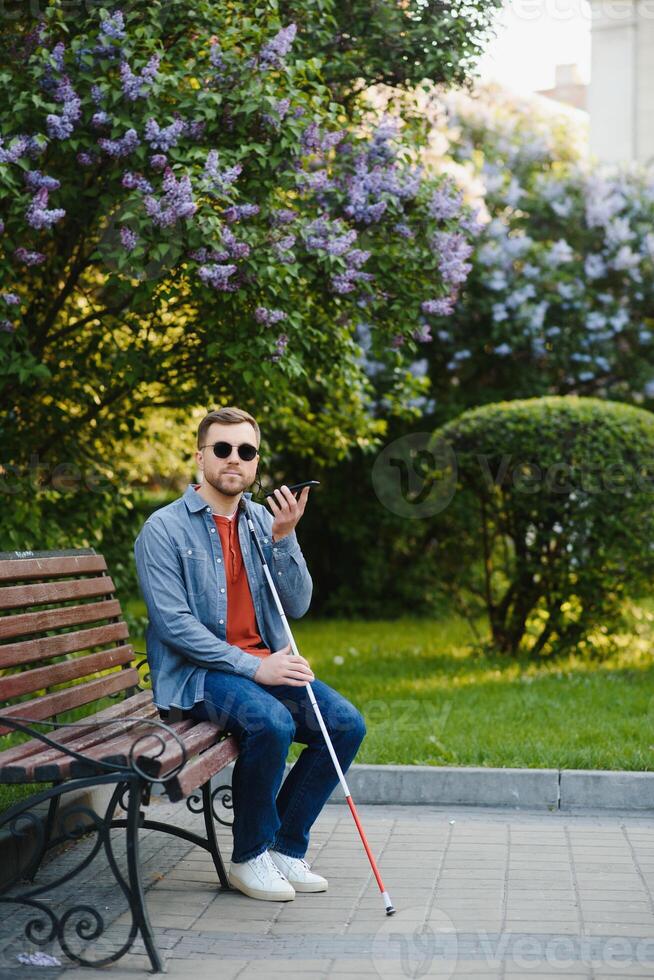 Visually impaired man with walking stick, sitting on bench in city park. Copy space photo