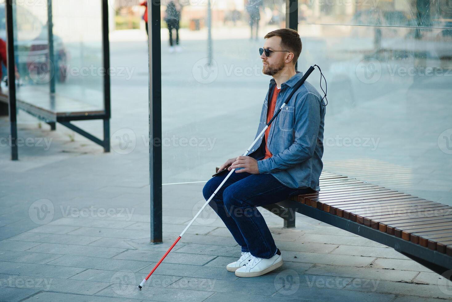 Visually impaired man with walking stick, sitting on bench in city park. Copy space photo
