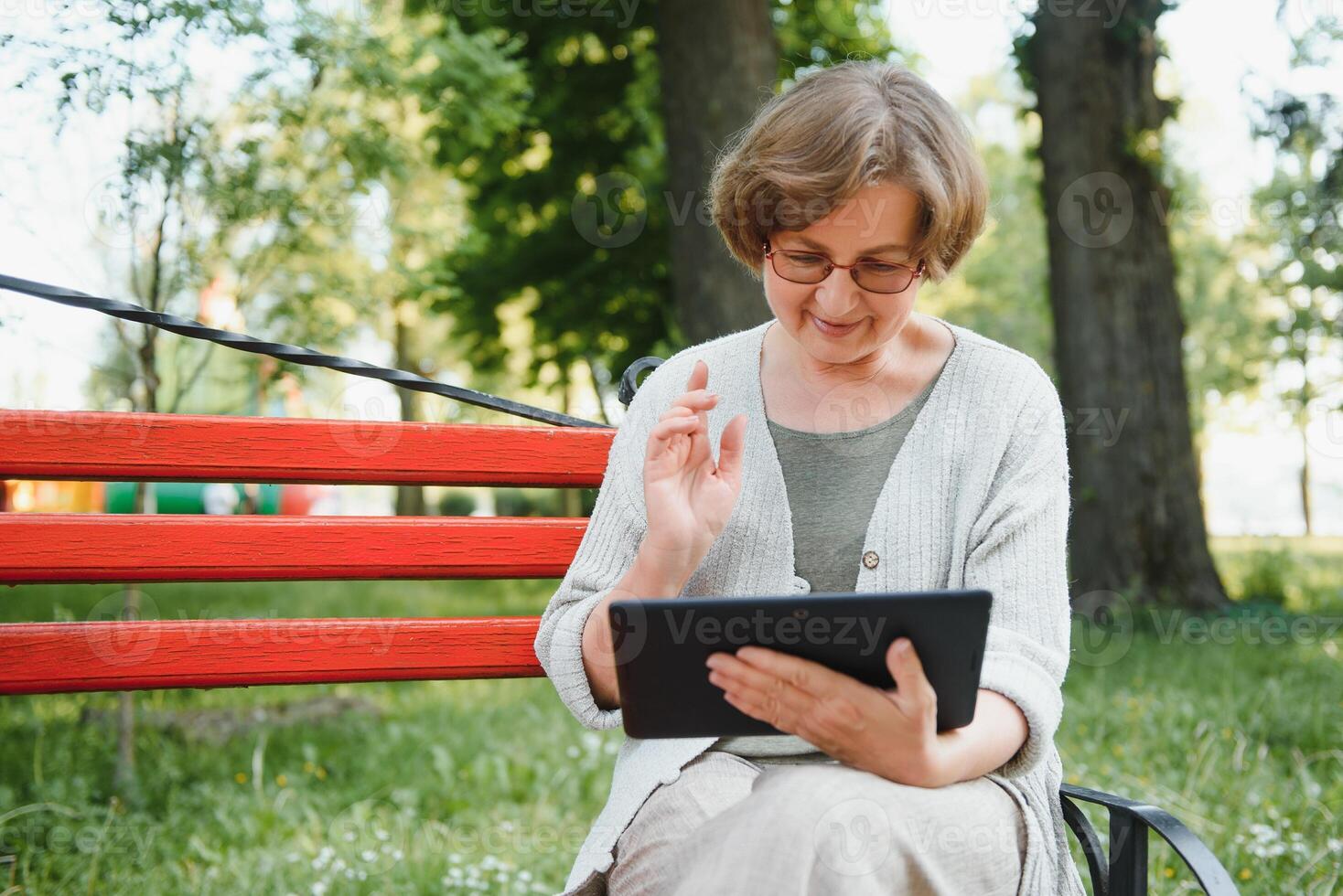 Positive confident old lady posing with tablet in park. Senior grey haired woman in casual sitting on park bench and using tablet. Wireless connection concept photo