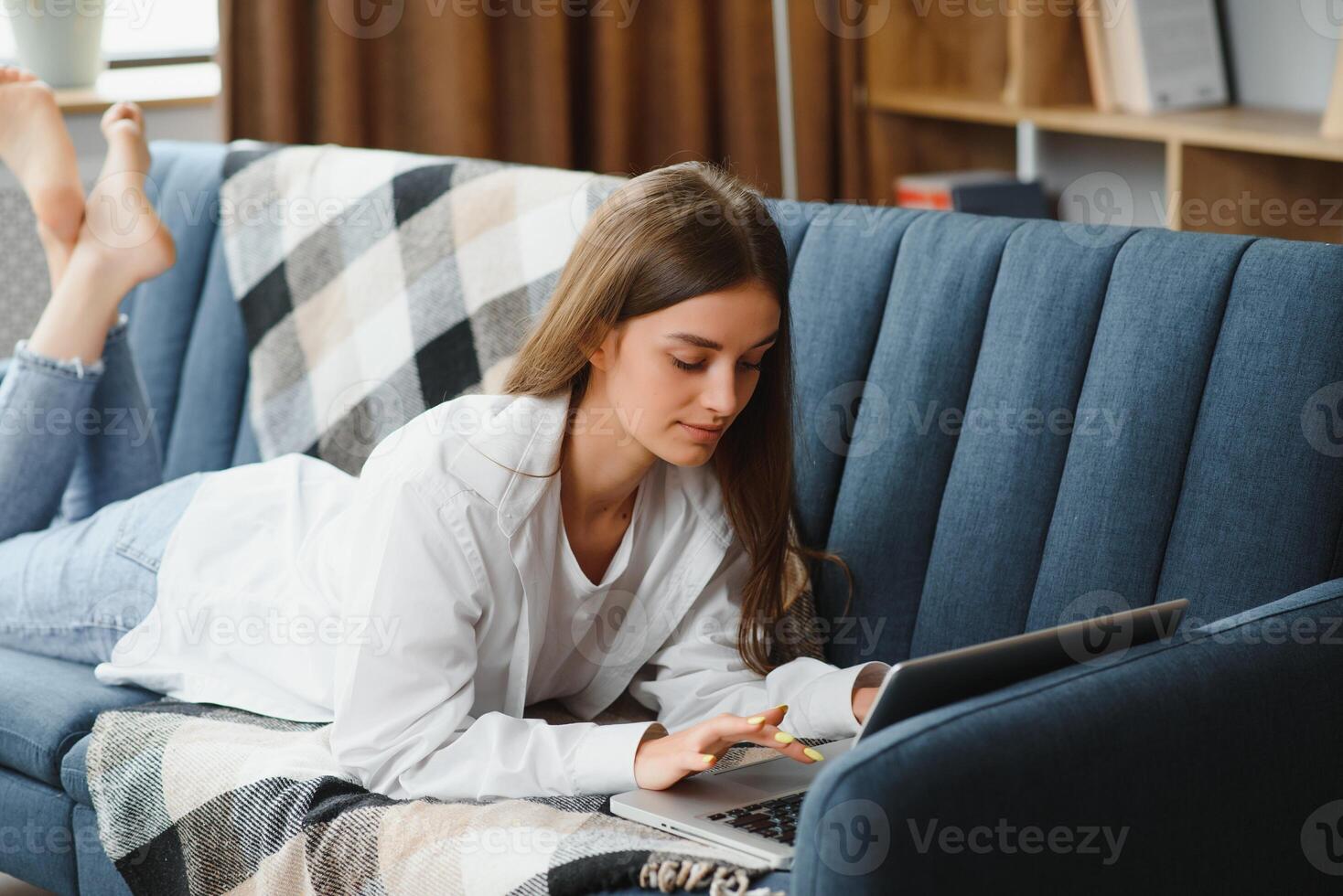 Smiling attractive young woman sitting on sofa using laptop communicating working online at home, happy teen girl typing on computer, enjoying writing blog or chatting with friends in social network photo